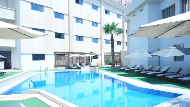 Una piscina con sillas de descanso y sombrillas, rodeada por un edificio de varios pisos y ventanas.