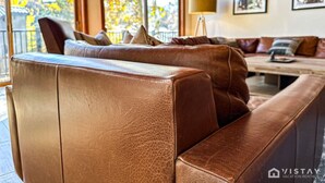 Ein braunes Ledersofa mit Kissen in einem gut beleuchteten Raum mit großen Fenstern, einer Lampe und gerahmten Bildern an der Wand.