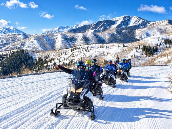 Un grupo de personas en motos de nieve en un sendero nevado, con montañas al fondo.