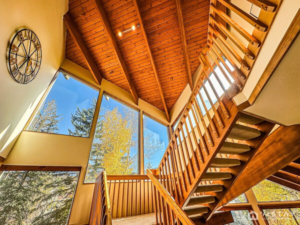 A wooden staircase with a clock on the wall, large windows, and a view of trees.