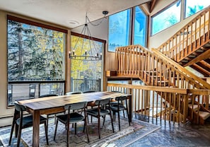 A dining area with a wooden table and chairs, large windows with a view of trees, and a wooden staircase. - Mountain Escape: Sauna & Fireplace (Park City)