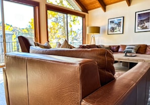 A brown leather sofa with cushions in a well-lit room with large windows, a lamp, and framed pictures on the wall. - Mountain Escape: Sauna & Fireplace (Park City)