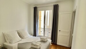 Un lit bien fait, avec des oreillers et une couverture, une table de chevet avec une lampe, une fenêtre avec des rideaux et un plancher en bois.