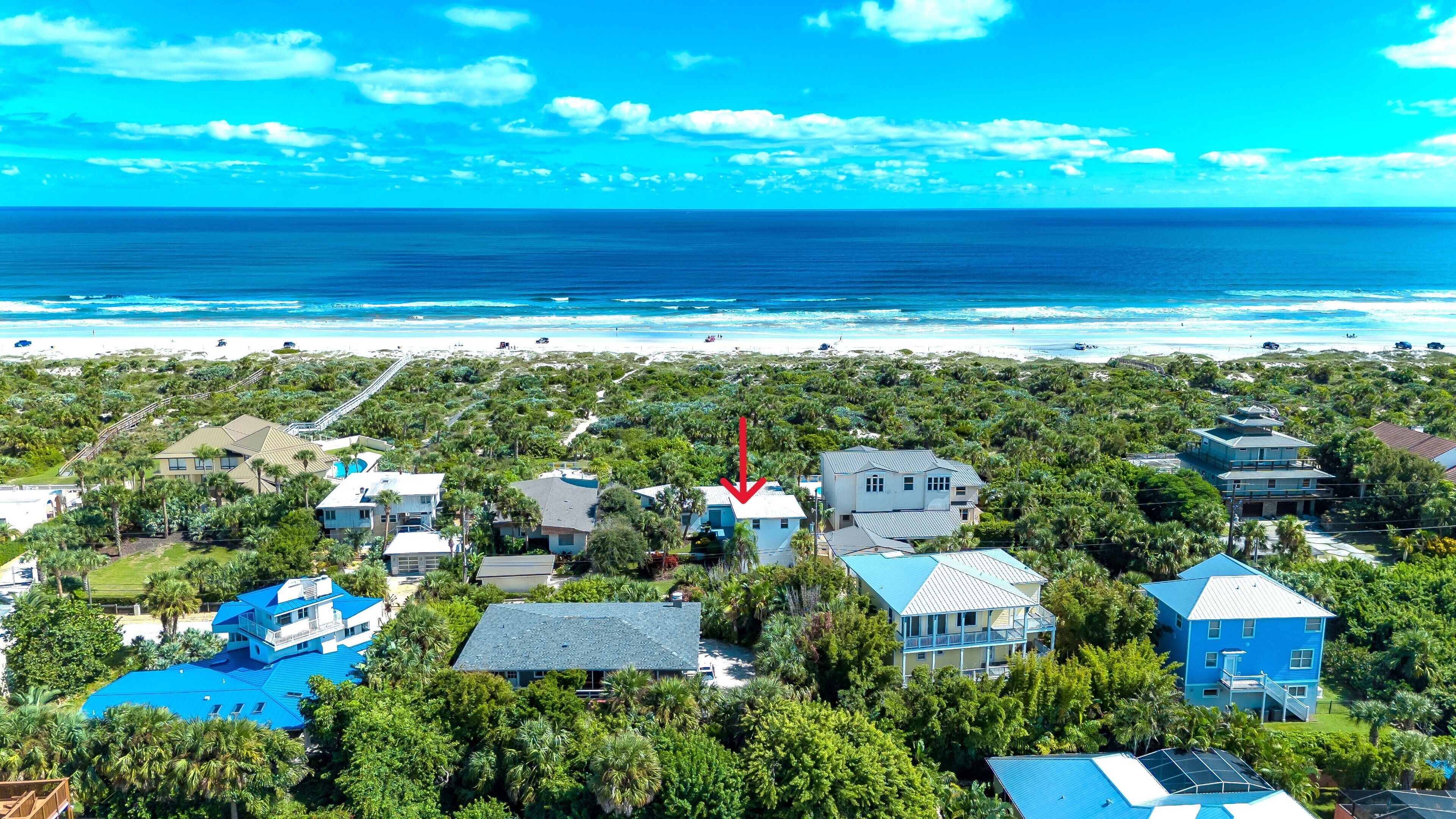 A coastal residential area with houses surrounded by dense vegetation, a beach in the background, and a clear blue sky.