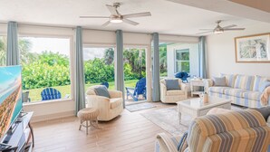 Un salon spacieux doté d’un grand téléviseur, de sièges confortables, de ventilateurs de plafond et offrant une vue sur une végétation luxuriante.
