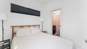Un dormitorio con cama, dos mesitas de noche, una mesilla auxiliar, una ventana con persianas y una puerta que da al baño.
