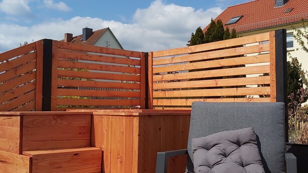 A wooden deck with a grey sofa and a table, a wooden fence, and a house with a red roof in the background. - Familienfreundliche Ferienwohnung in Wernigerode, mit Eigenem Garten (Wernigerode)