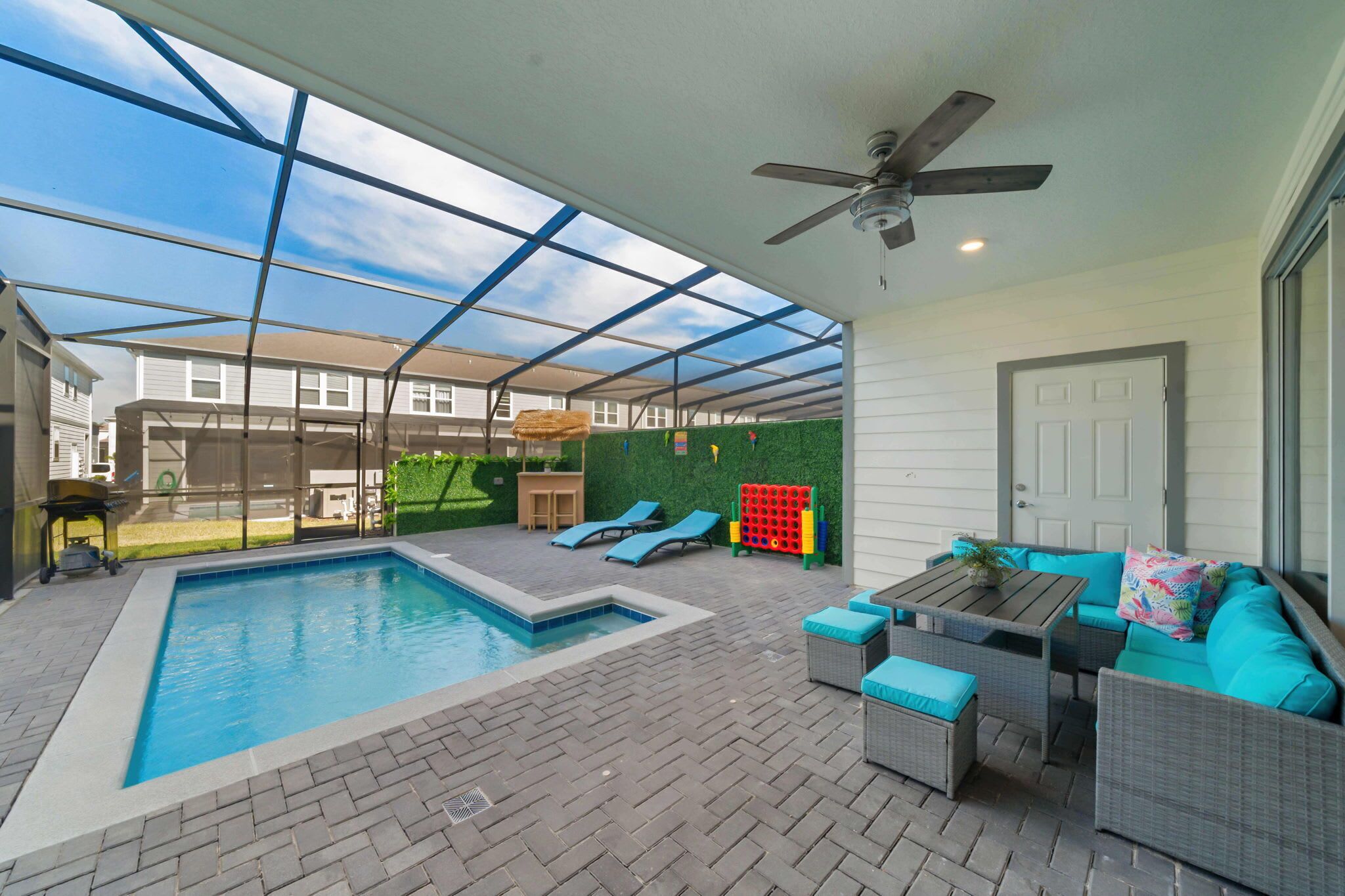 A covered outdoor swimming pool with lounge chairs and a table, surrounded by a patio area with a ceiling fan and a view of neighboring houses.