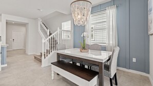 A dining area with a white table, dark wooden bench, wicker chairs, a chandelier, and a staircase.