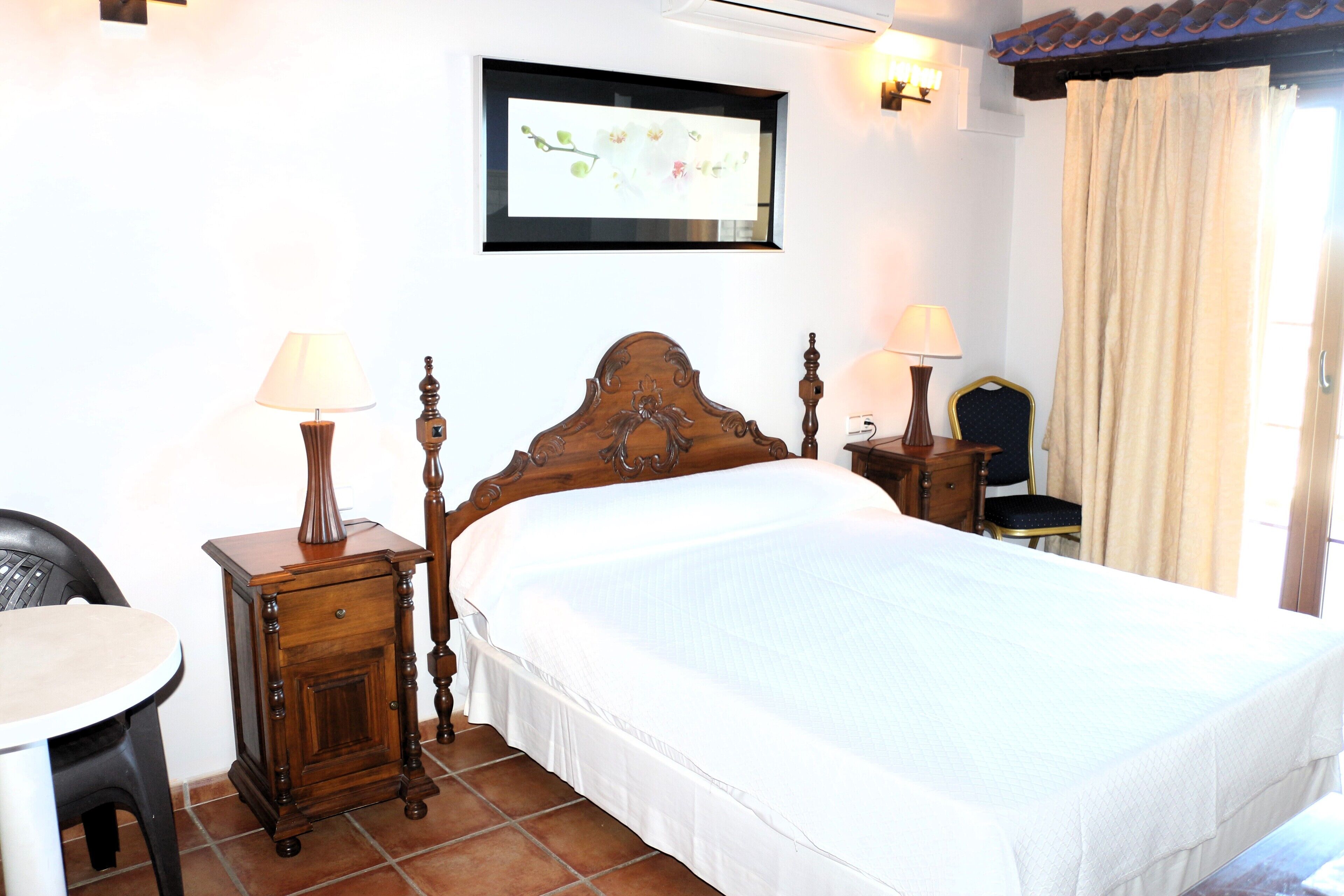 Un dormitorio ordenado con una cama de madera, dos mesitas de noche con lámparas, una silla e imagen enmarcada en la pared.