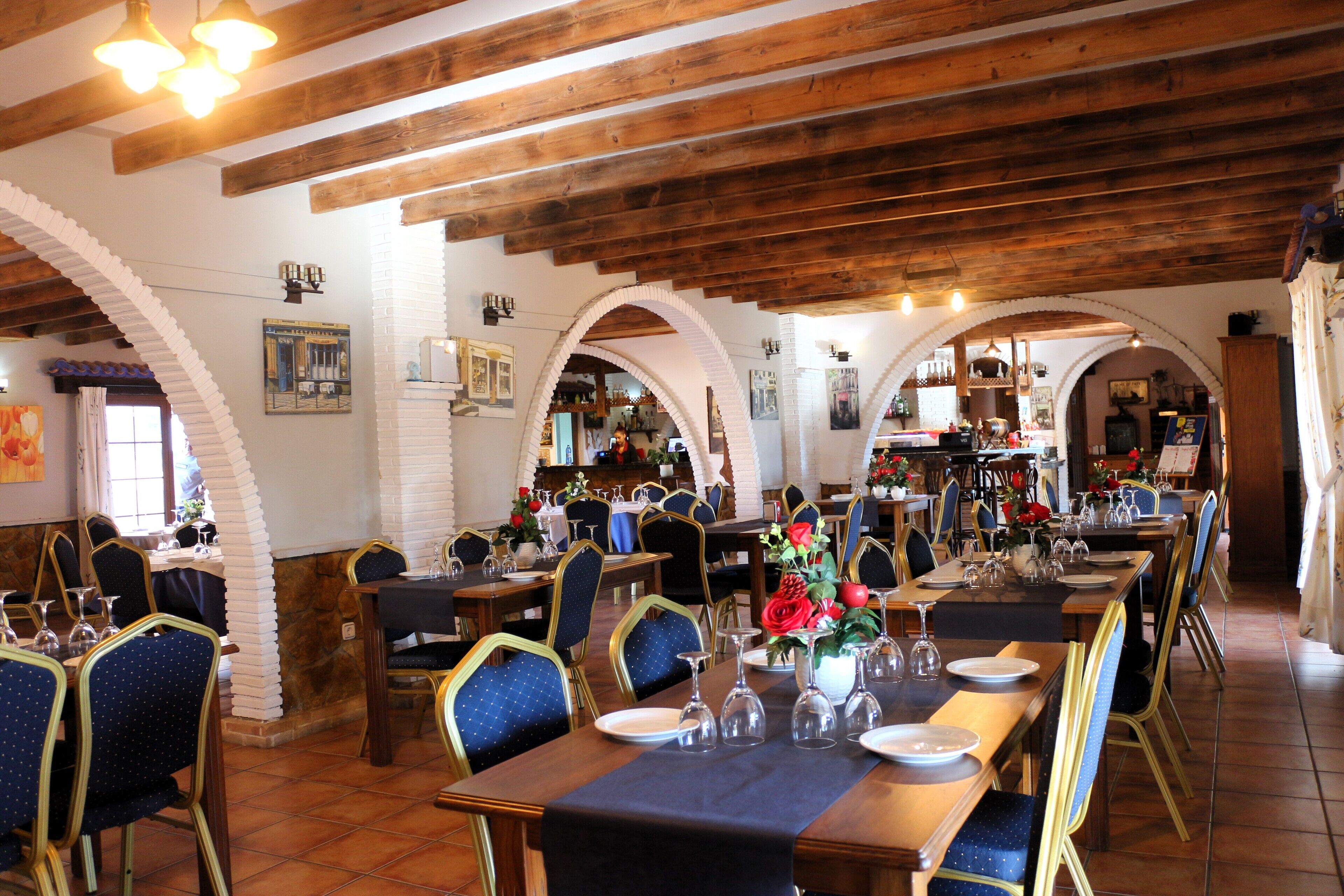 Een restaurant met houten balken, gewelfde plafonds en blauwe beklede stoelen. Tafels zijn gedekt met borden, glazen en bloemen.