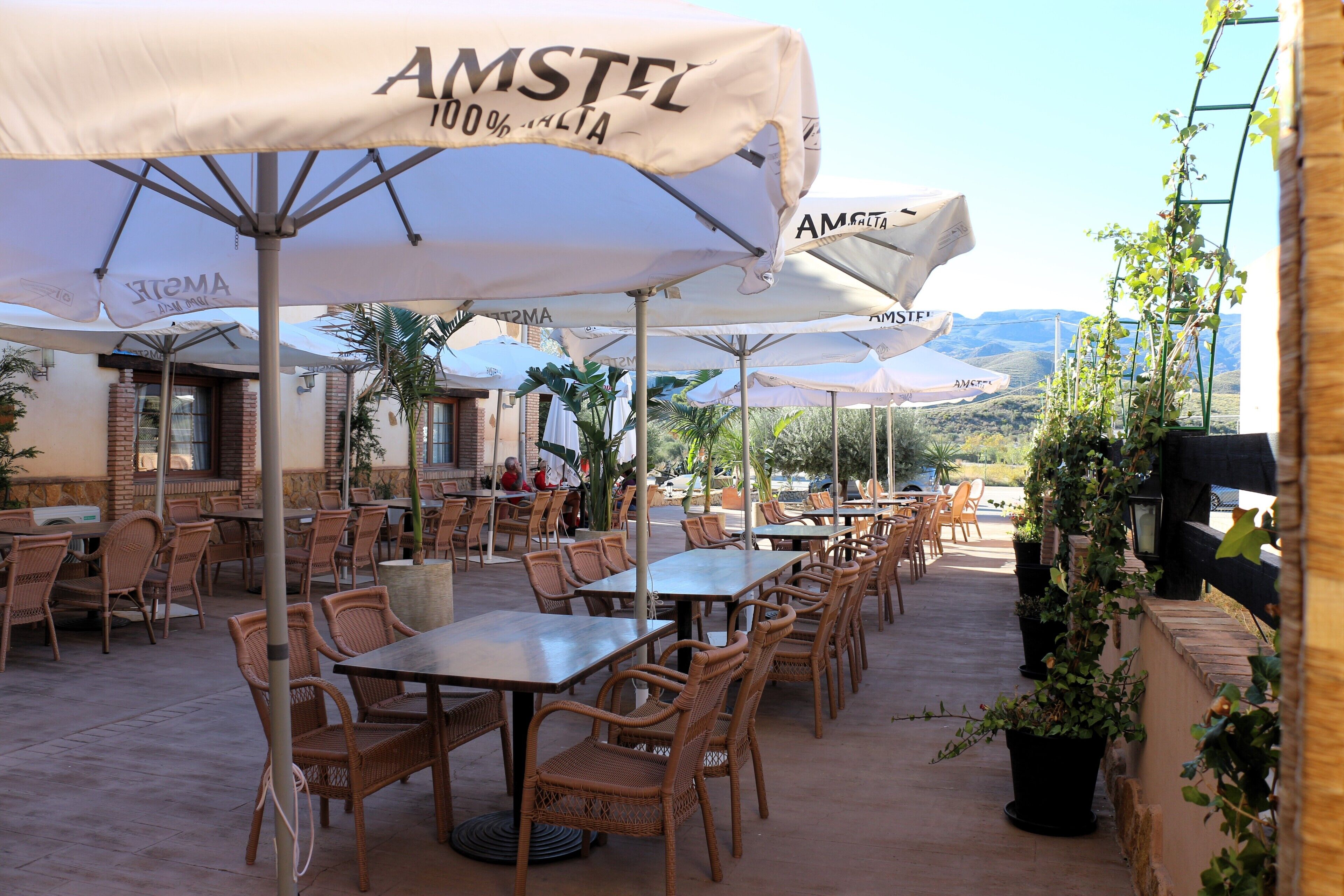 Un caffè all'aperto con tavoli e sedie in vimini, riparato da ombrelloni bianchi con marchio 'AMSTEL', sullo sfondo di dolci colline.