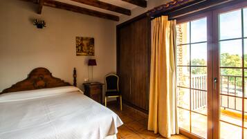 A bedroom with a large bed, a chair, a nightstand, a lamp, a painting on the wall, and a balcony with a view.