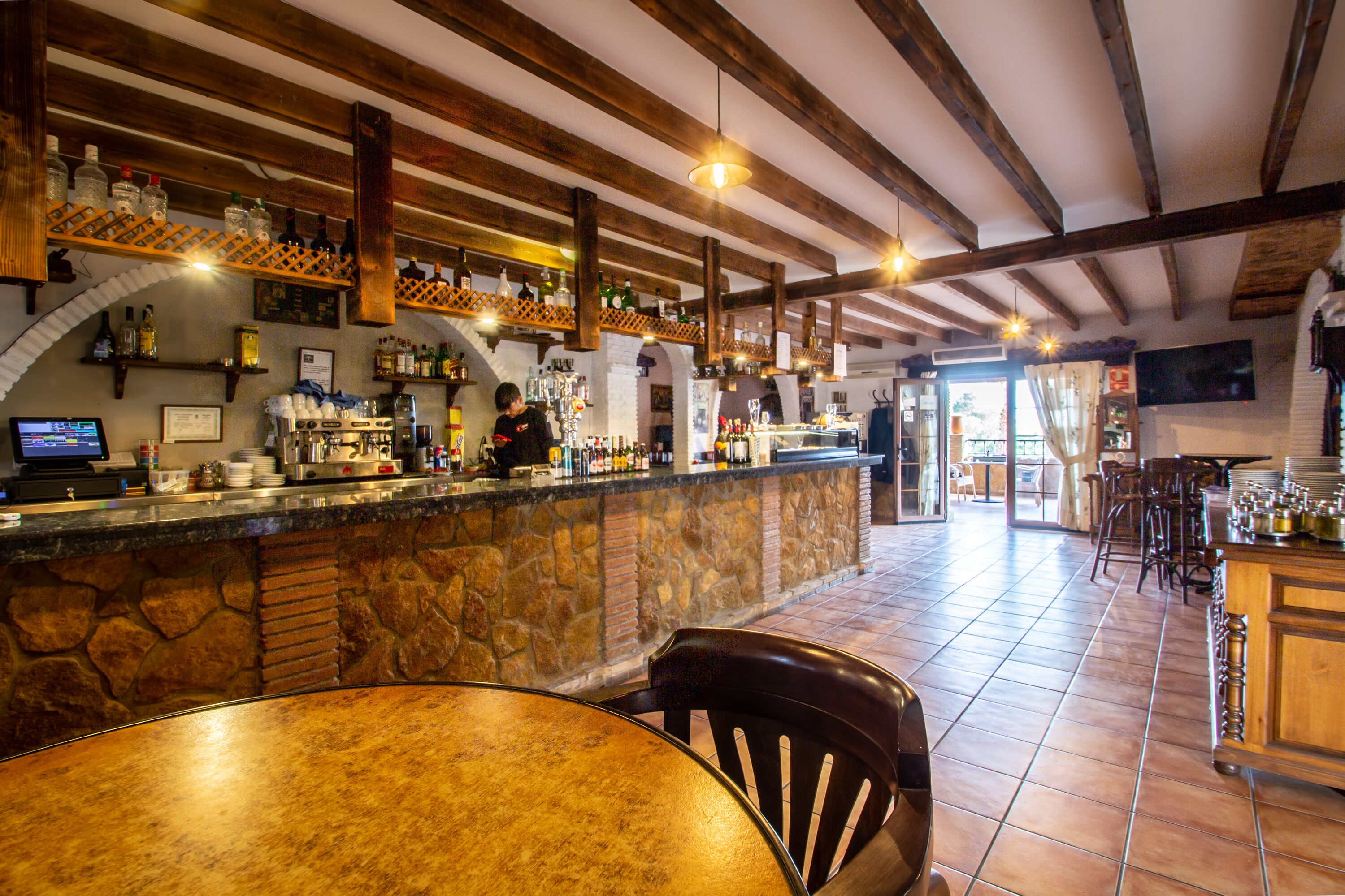 A bar area with a stone counter, wooden beams, and a variety of bottles and glasses.