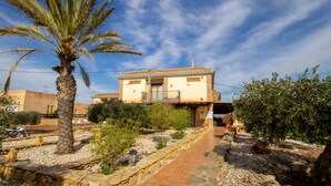 Una casa de dos pisos con un patio empedrado, una palmera y un jardín con diversas plantas.