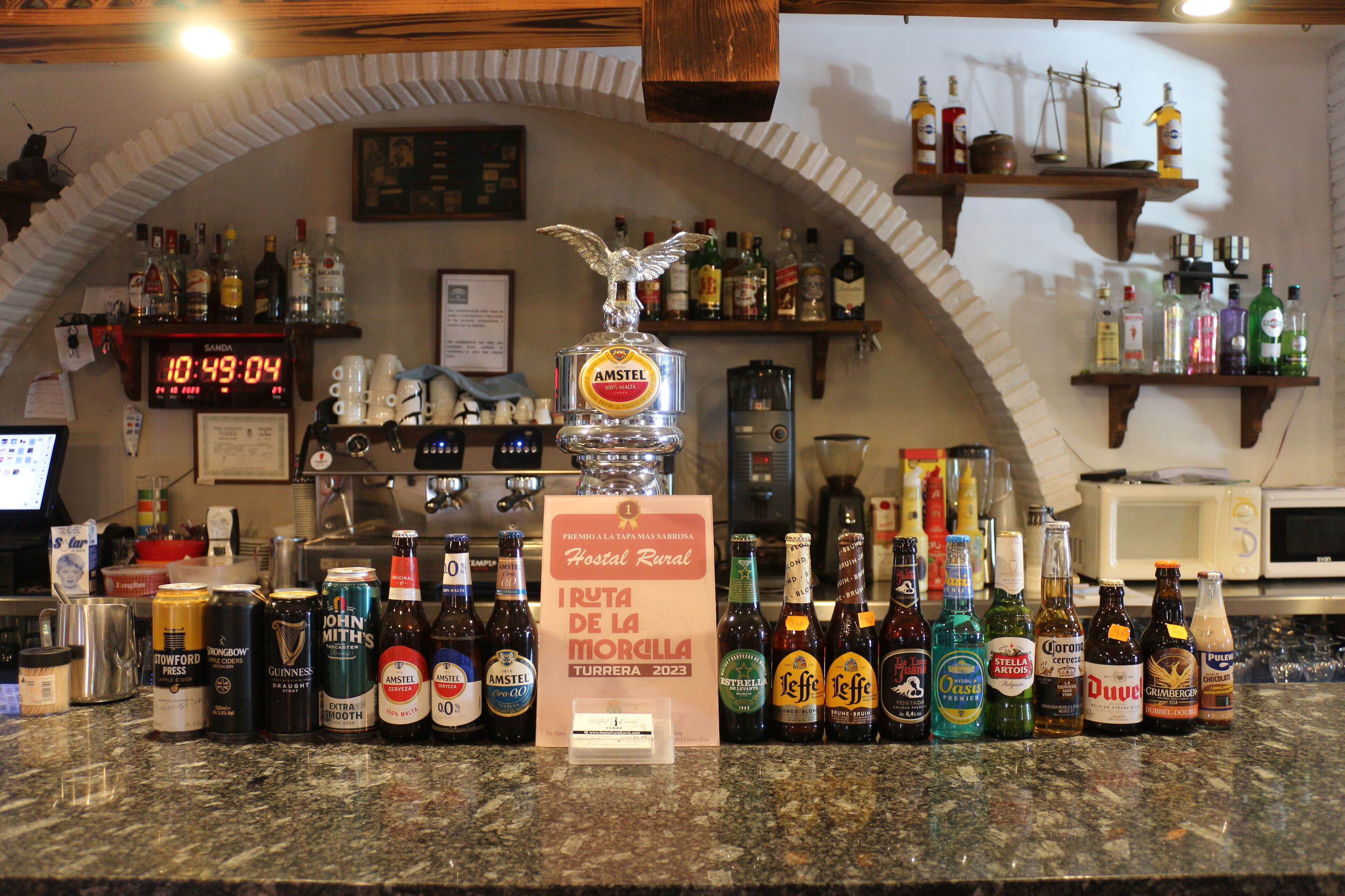 A bar area with a variety of liquor bottles, a clock displaying 10:49:04, and a sign for 'Hostal Rural'.