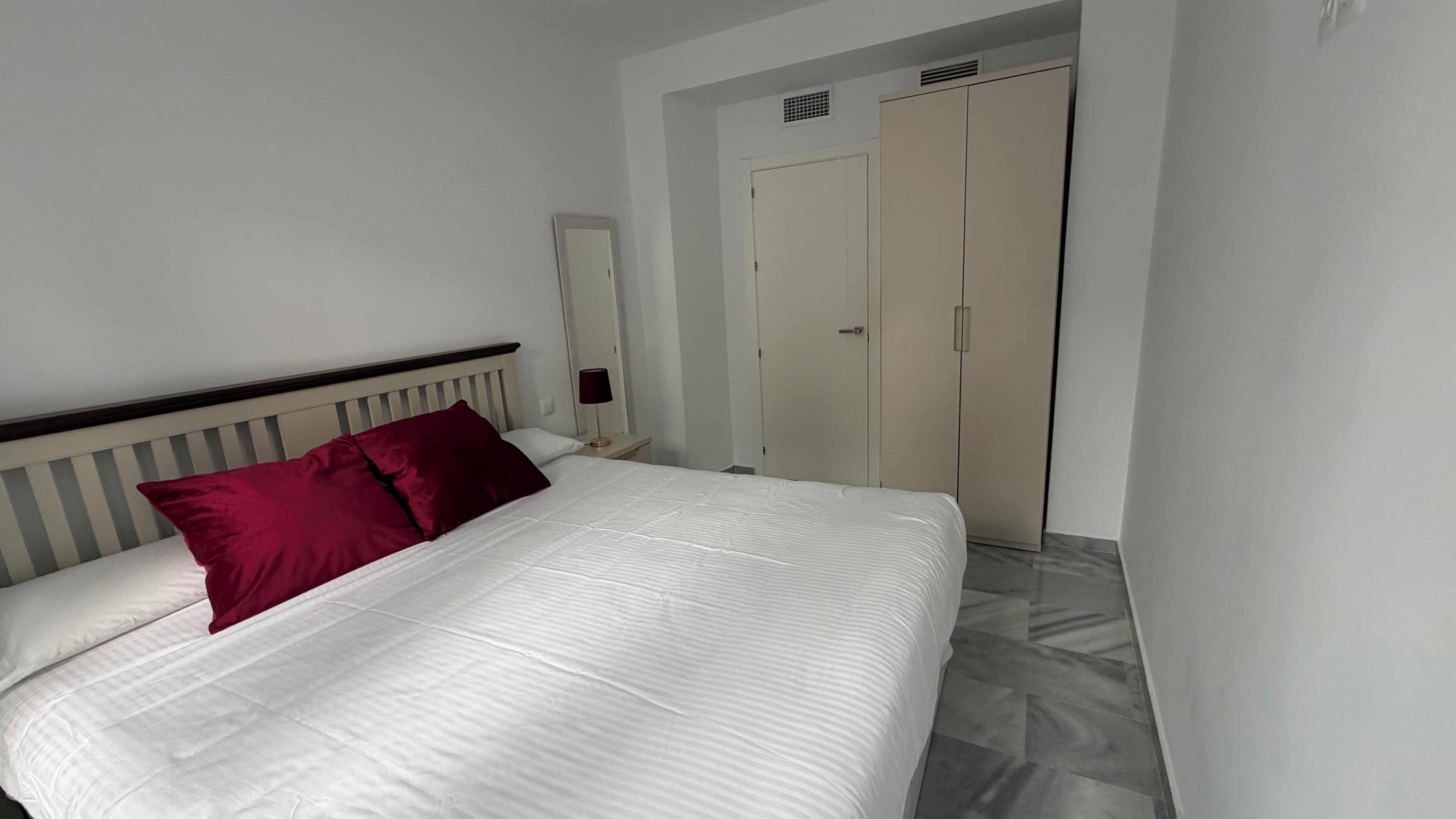 A bed with white bedding and a red pillow, a bedside table with a lamp, and a wardrobe with a door.