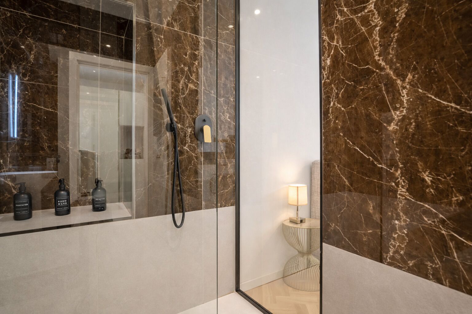 A modern bathroom with a glass shower enclosure, marble walls, and a small bedside table with a lamp.