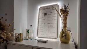 Une pancarte d’accueil indiquant les règles de la maison, un vase avec des fleurs séchées, un verre et un petit appareil posés sur un comptoir blanc.