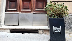 Une grande porte double en bois, surmontée d’une petite boîte aux lettres et, au sol, un panneau noir portant l’inscription « Milano Design District ».