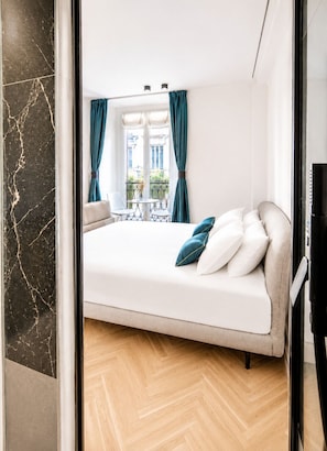Une chambre à coucher avec un lit, une fenêtre avec des rideaux, un balcon et une vue sur des immeubles.