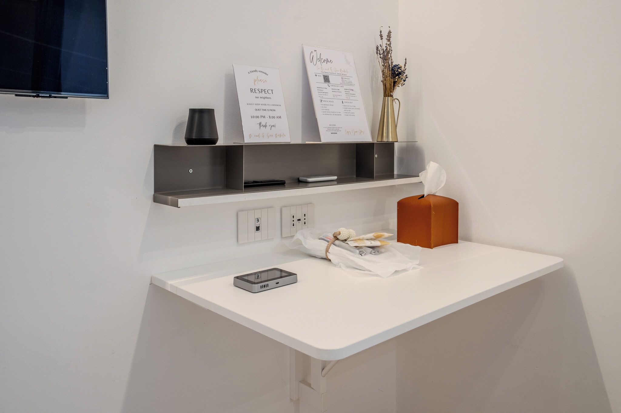 A reception desk with a flat-screen TV, two welcome signs, a vase with dried flowers, and a tissue box.