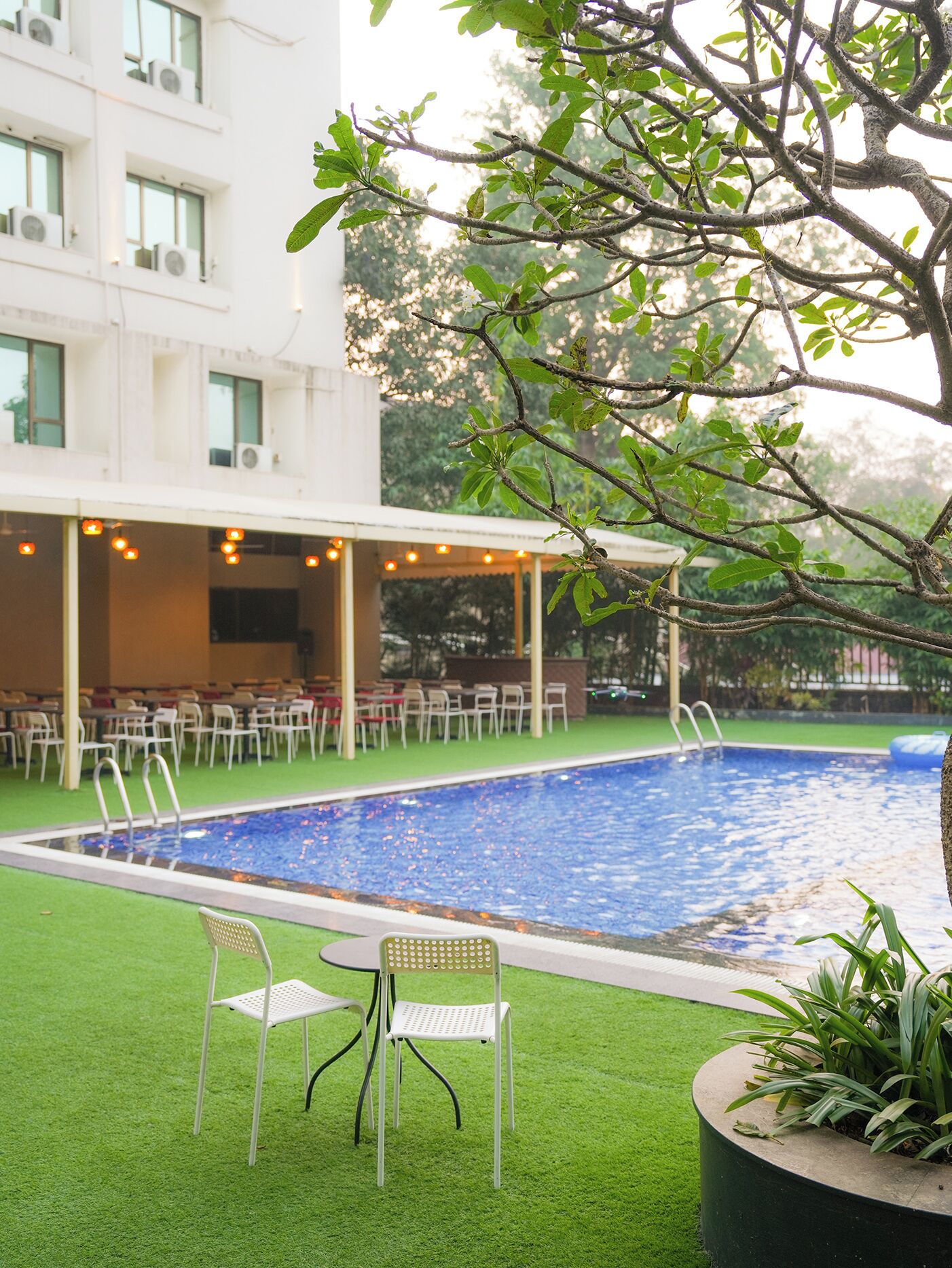 A swimming pool with a covered seating area, surrounded by greenery and a building with multiple windows.
