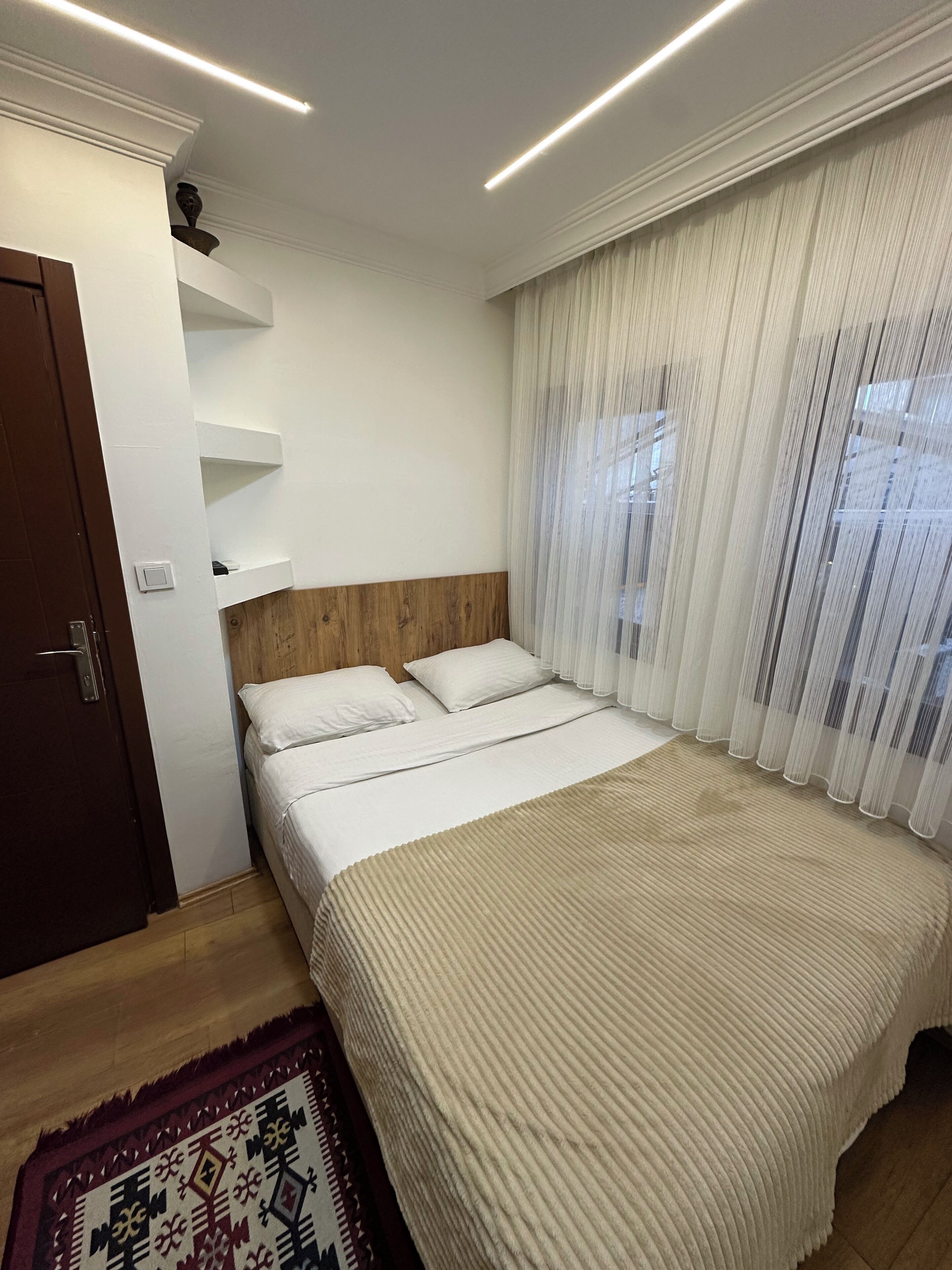 A bedroom with a bed, a wooden headboard, a white wall, a door, and a window with sheer curtains.