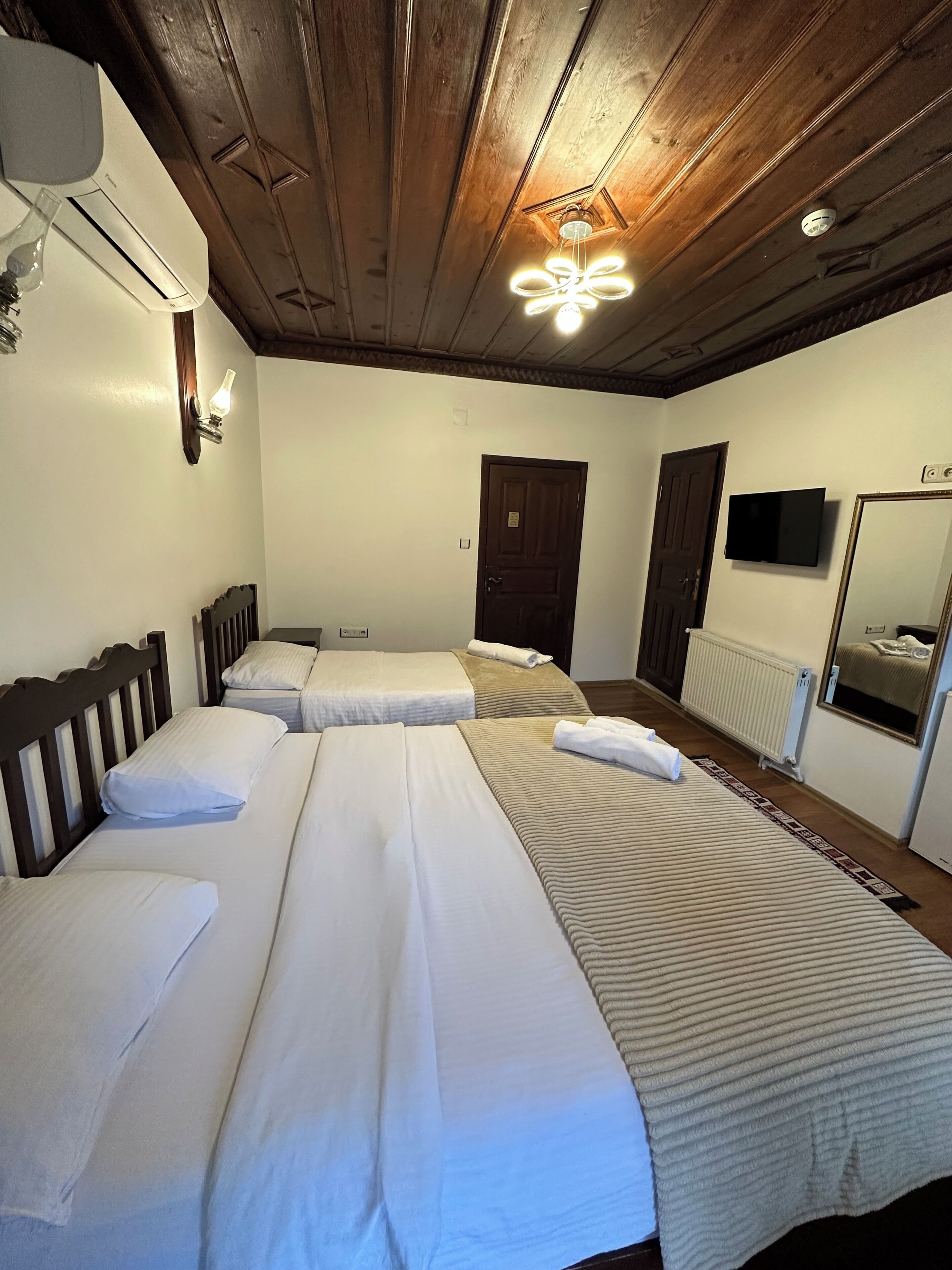A room with two beds, a wooden ceiling, a wall-mounted TV, and a radiator.