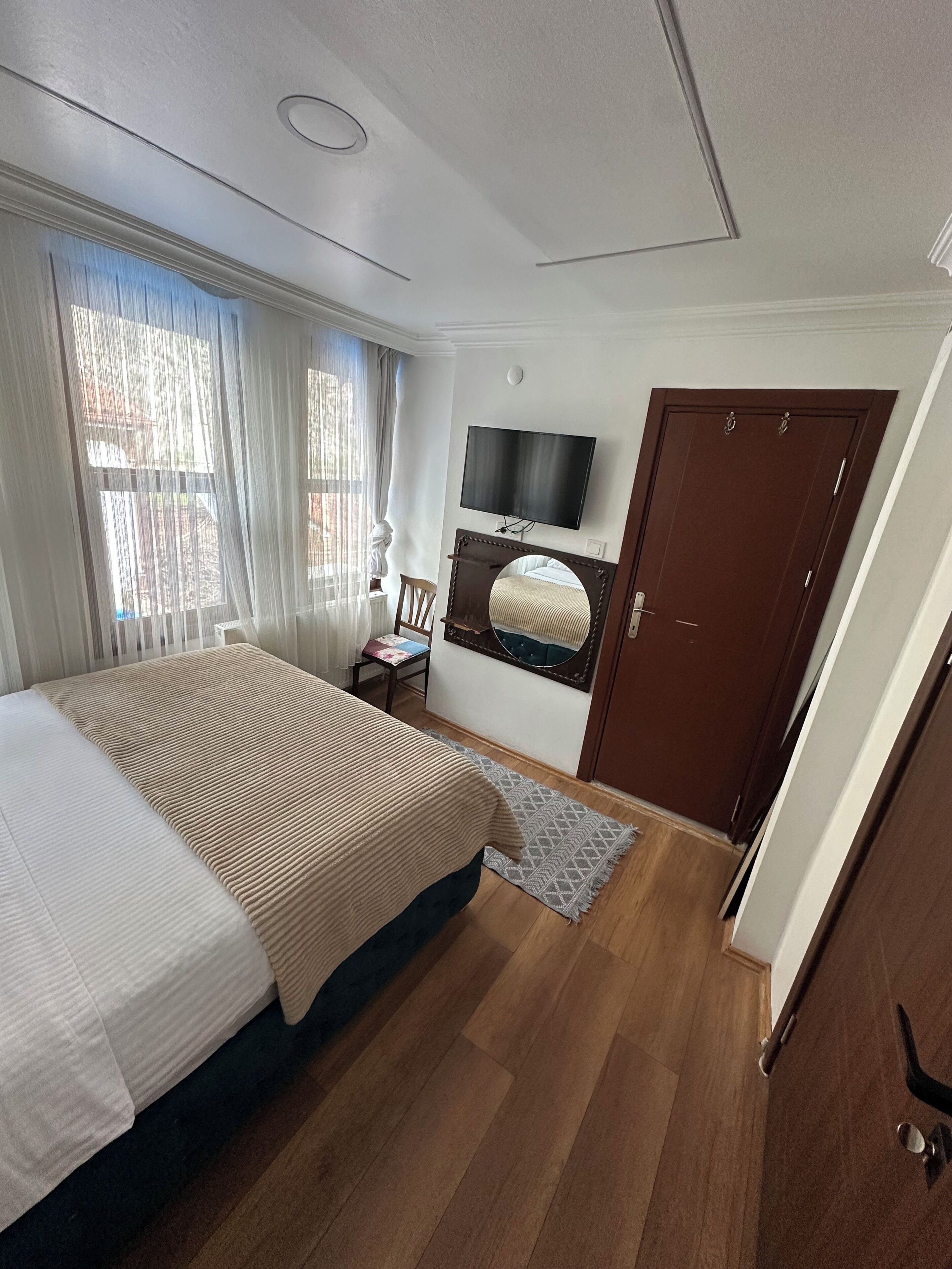 A bedroom with a bed, a TV, a wooden door, and a window with curtains.