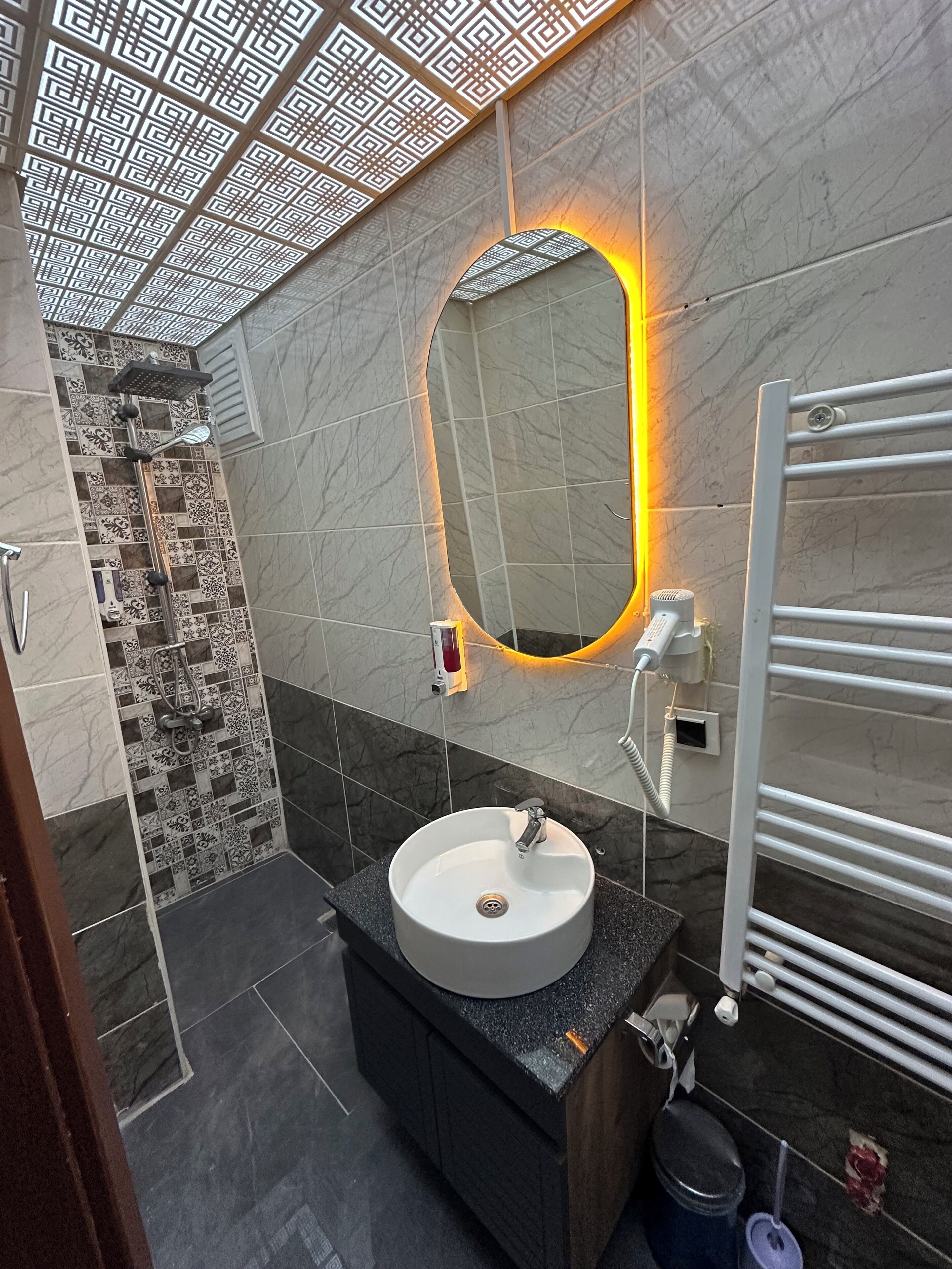 Un baño moderno con un espejo ovalado grande, un lavabo redondo, una ducha con diseño geométrico en el techo y un perchero para toallas.