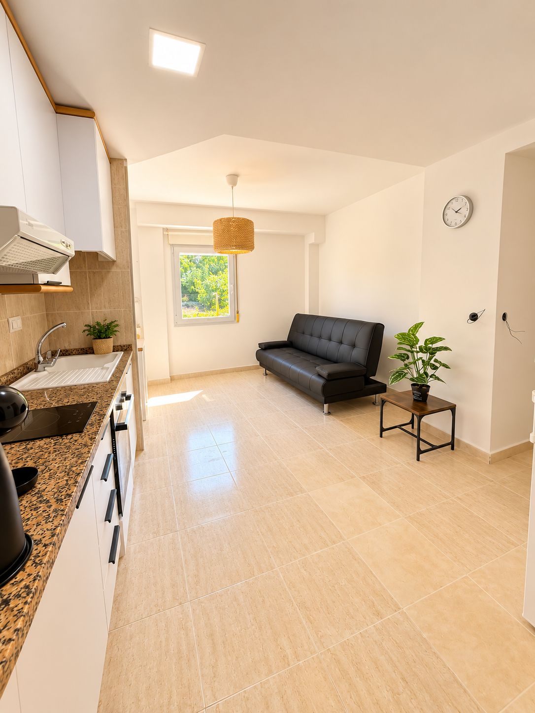 A modern kitchen with a built-in oven, a sink, and a stove. There is a black leather sofa and a small wooden table with a plant. A window provides natural light, and a clock is on the wall.