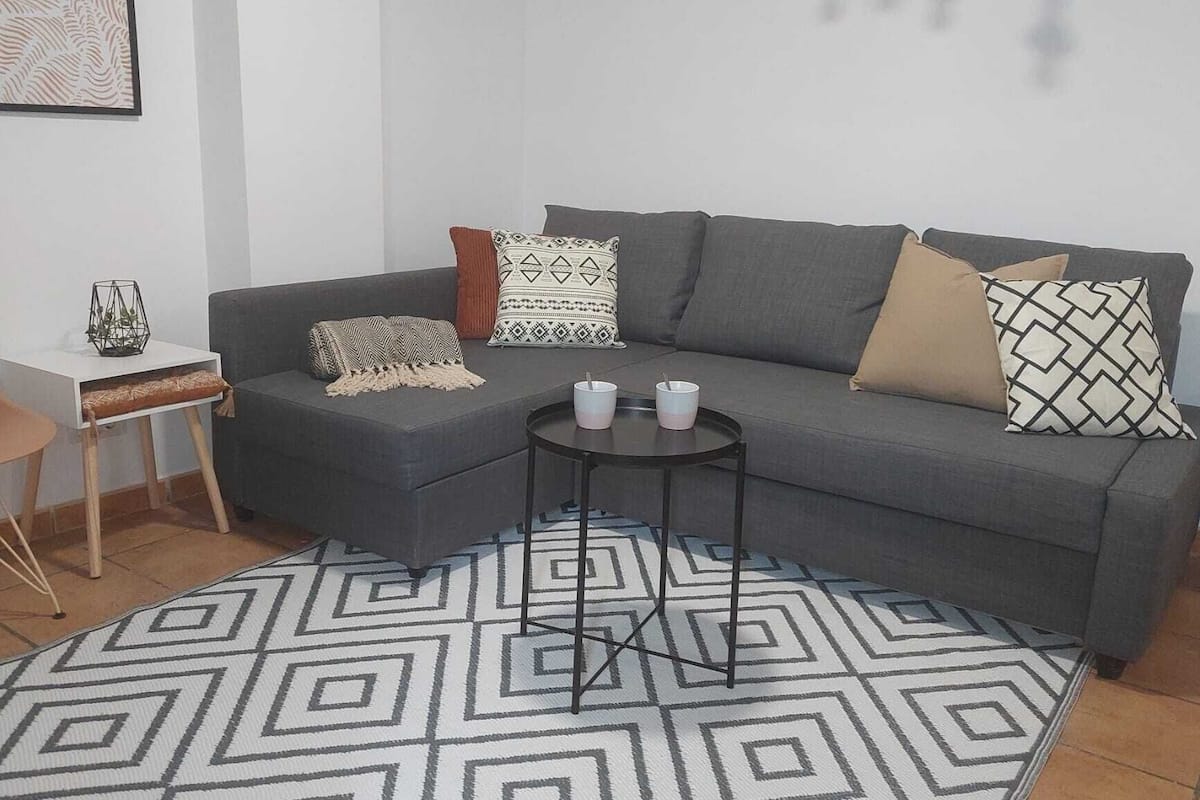 Ett modernt vardagsrum med en grå soffa, mönstrade kuddar, ett litet sidobord, ett soffbord och en geometrisk matta.