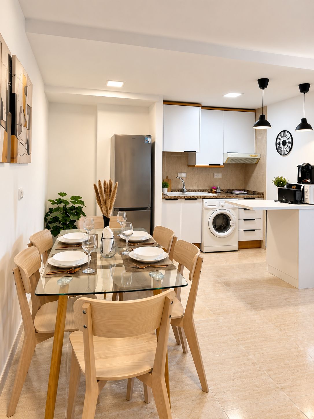 A modern kitchen with white cabinets, a stainless steel refrigerator, a washing machine, and a dining area with a glass table and wooden chairs.