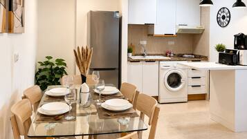 Una cocina moderna con armarios blancos, un refrigerador de acero inoxidable, una lavadora y un comedor con una mesa de vidrio y sillas de madera.