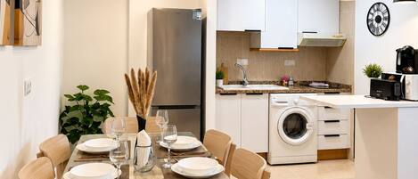 A modern kitchen with white cabinets, a stainless steel refrigerator, a washing machine, and a dining area with a glass table and wooden chairs.