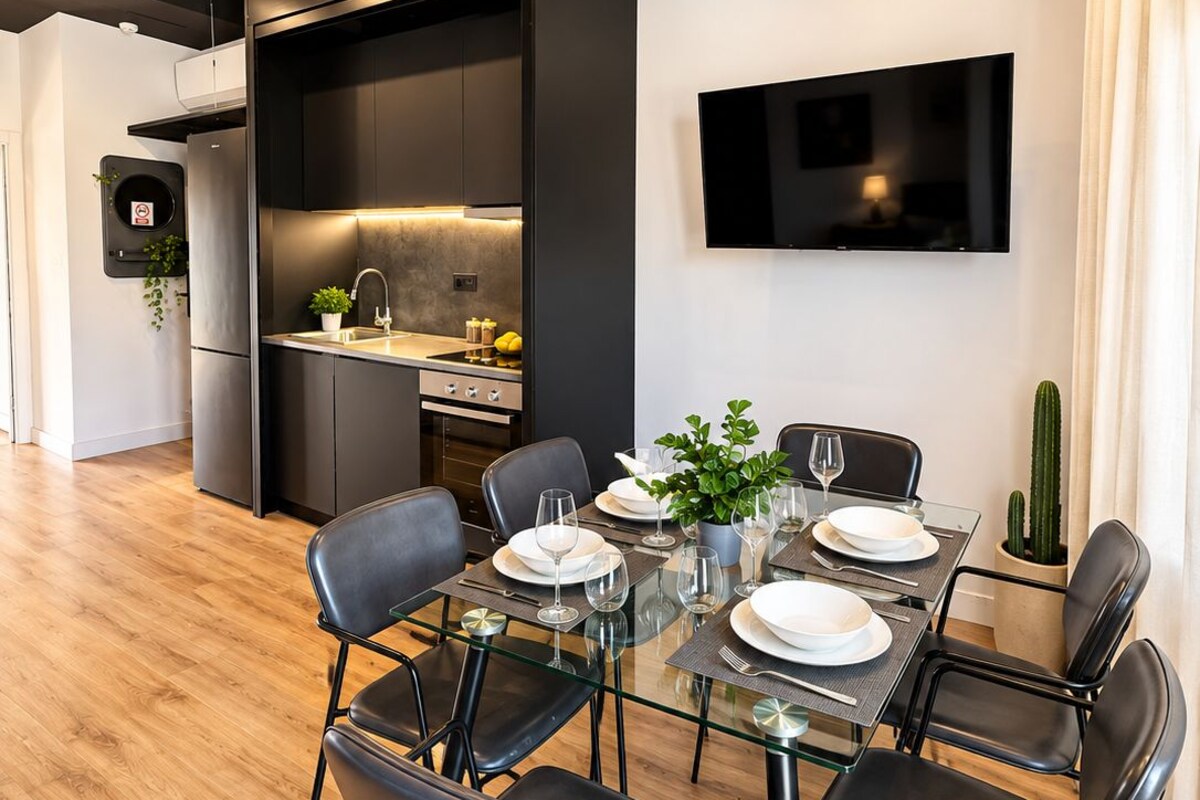 A modern dining area with a glass table, black chairs, and a kitchen in the background.