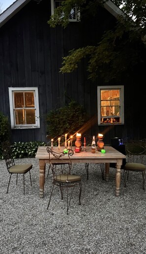 Et udendørs spiseområde med et træbord og stole, omgivet af grus, foran en mørk træbygning med vinduer.