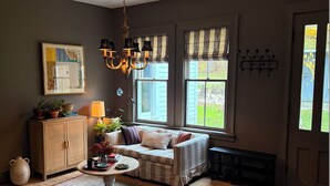 A cozy living room with a sofa, a coffee table, and a chandelier. There are windows with curtains, a cabinet, and a rug on the floor.