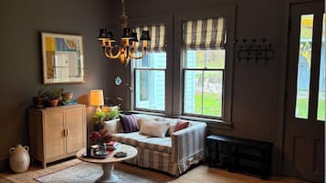 A cozy living room with a sofa, a coffee table, and a chandelier. There are windows with curtains, a cabinet, and a rug on the floor.