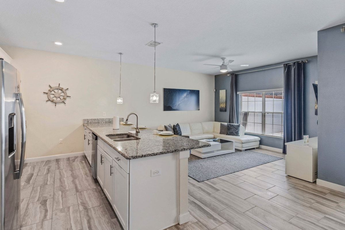 A modern kitchen with a central island, built-in appliances, pendant lighting, and a dining area with a sofa and a coffee table.