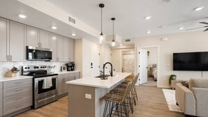 A modern kitchen with stainless steel appliances, a central island with bar stools, and a flat-screen TV in the living area. - Hummingbird Retreat 2 Bedroom Condo (St. George)