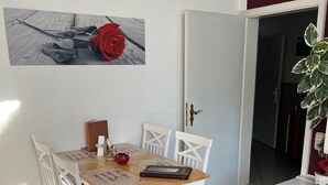 Uma área de refeições com mesa de madeira e cadeiras brancas, uma decoração floral vermelha e uma porta que dá para outro cômodo.