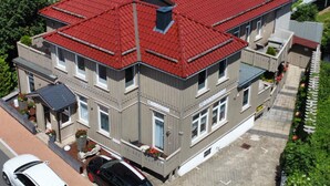 Um edifício de dois andares com telhado vermelho, várias janelas e uma pequena estrutura anexa com plantas em vasos.