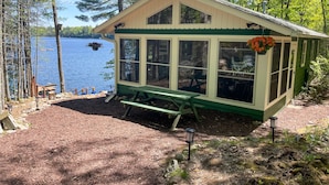 A small, single-story cabin with large windows, a green bench, and a hanging flower basket.