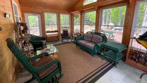 A cozy cabin interior with wooden walls, large windows, wicker furniture, and a view of the forest.