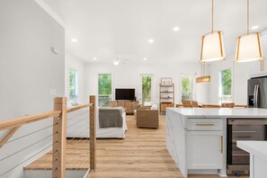 Un intérieur moderne comprenant une cuisine, un coin repas et un salon, avec des planchers en bois, des murs blancs et un éclairage suspendu.