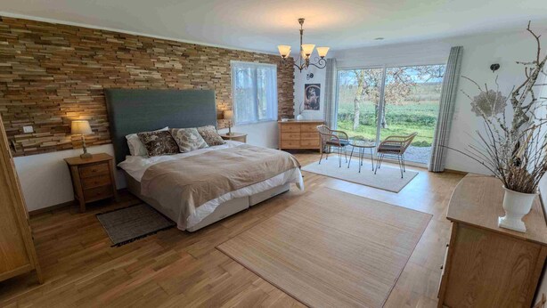 Ein Schlafzimmer mit einem Holzkopfteil, einem Bett mit beiger Bettdecke, einem Nachttisch, einer Kommode und Blick ins Freie durch ein großes Fenster.