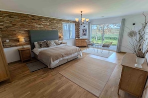 A bedroom with a wooden headboard, a bed with a beige comforter, a nightstand, a dresser, and a view of the outdoors through a large window. - Chez Phil, Chambres D'hôte, Chambre Thème Bois (La Pomarède)