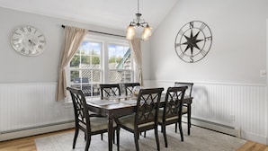 A dining room with a table set for four, surrounded by dark wooden chairs with cushioned seats. There are two wall clocks, one with Roman numerals and the other with a compass design. A chandelier hangs from the ceiling, and a window with curtains is visible in the background. - 3bed, 2bath Hamptons Home, Close to Incredible Beaches and Restaurants! (Hampton Bays)